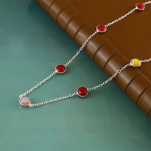 May include: A silver necklace with a delicate chain design. The necklace features several round, bezel-set gemstones in shades of red and yellow. The necklace is displayed on a teal surface with a brown leather background.