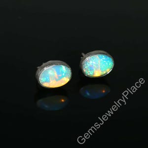 May include: A pair of oval opal stud earrings. The opals display a vibrant play of color, with hues of blue, green, and yellow. The earrings are set in a silver-toned metal. The background is black.