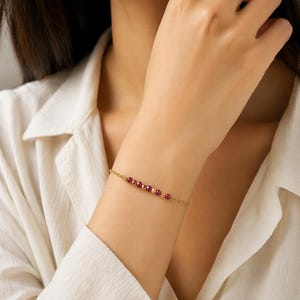 Könnte beinhalten: Ein zartes goldenes Armband mit einer Reihe kleiner, facettierter roter Perlen, die mit goldfarbenen Perlen durchsetzt sind. Das Armband wird an einem Handgelenk getragen und zeigt sein schlichtes, aber elegantes Design. Der Schmuck ist vor einem weißen Hemd platziert.