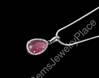 May include: A silver necklace with a pendant featuring a pear-shaped, pink gemstone surrounded by small, clear stones. The pendant hangs from a delicate silver chain against a black background.