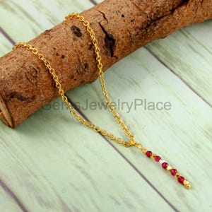 Pode incluir: Um colar dourado com uma corrente delicada e um pingente vertical. O pingente apresenta uma série de pequenas pedras preciosas vermelhas redondas e algumas pedras brancas. O colar é exibido em um pedaço de madeira.