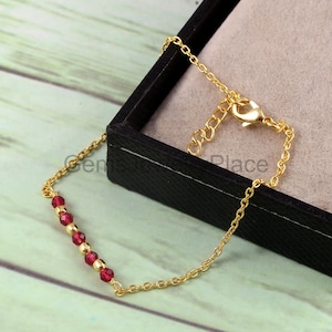 Pode incluir: Uma pulseira de corrente dourada com uma série de pequenas contas redondas de cor vermelho escuro. A pulseira é exibida em uma caixa de joias preta com interior cinza claro. O fecho é visível.