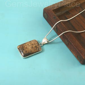 Op de afbeelding: Een rechthoekige hanger met een bruine steen en zilverkleurige rand hangt aan een zilveren ketting. De hanger is geplaatst tegen een blauwe achtergrond en een houten oppervlak. De sieraden worden in een studio gepresenteerd.