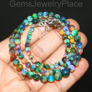 May include: A multi-strand necklace composed of iridescent opal beads in shades of green, blue, orange, and brown. The necklace is secured with a silver-colored clasp. The text "GemsJewelryPlace" is visible at the top.