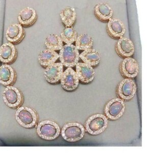 May include: A gold-toned jewelry set featuring an opal pendant, matching earrings, and a bracelet. The opal stones have a colorful, iridescent appearance and are surrounded by small, clear gemstones. The set is displayed on a gray surface.