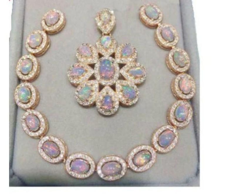 May include: A gold-toned jewelry set featuring an opal pendant, matching earrings, and a bracelet. The opal stones have a colorful, iridescent appearance and are surrounded by small, clear gemstones. The set is displayed on a gray surface.