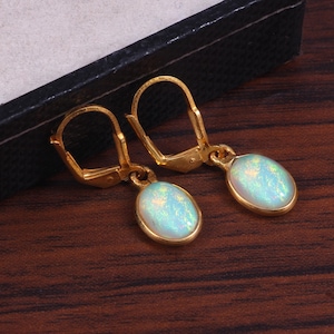 May include: A pair of gold-colored leverback earrings. Each earring features an oval, iridescent opal gemstone set in a gold bezel. The gemstones display a mix of pale blue, green, and yellow hues. The earrings are displayed against a dark background.