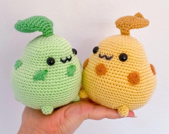 May include: Two crocheted plushies, one green with a leaf on top and one yellow with a brown leaf on top. Both plushies have black eyes and a smiling mouth.