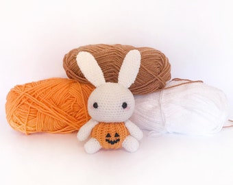 May include: A white crocheted bunny wearing a pumpkin costume sits in front of three balls of yarn: orange, brown, and white.
