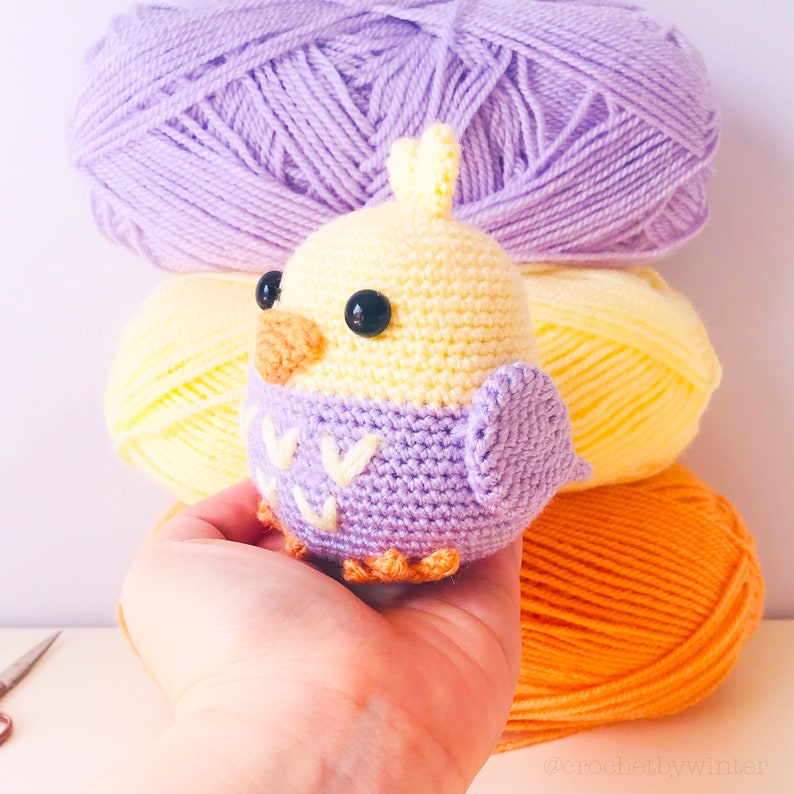 May include: A crocheted yellow bird with a purple vest and black eyes. The bird is sitting on a hand, with balls of yarn in the background.
