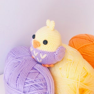 May include: A crocheted yellow bird with purple wings and a black eye, sitting on a pile of purple, yellow, and orange yarn.