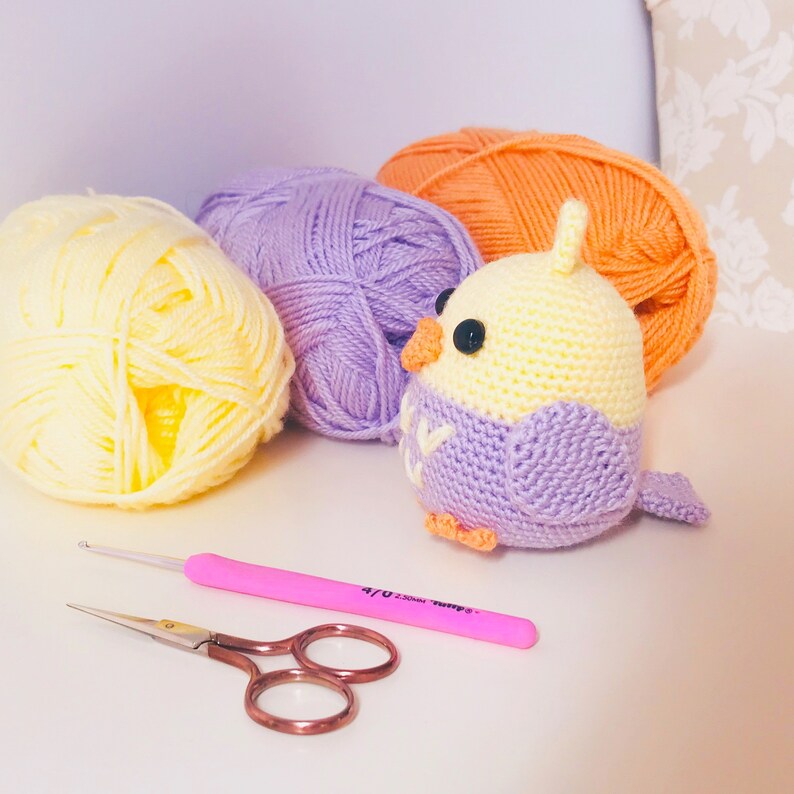 May include: A crocheted bird toy with a yellow body and purple wings, sitting on a table with three balls of yarn in yellow, purple, and orange.