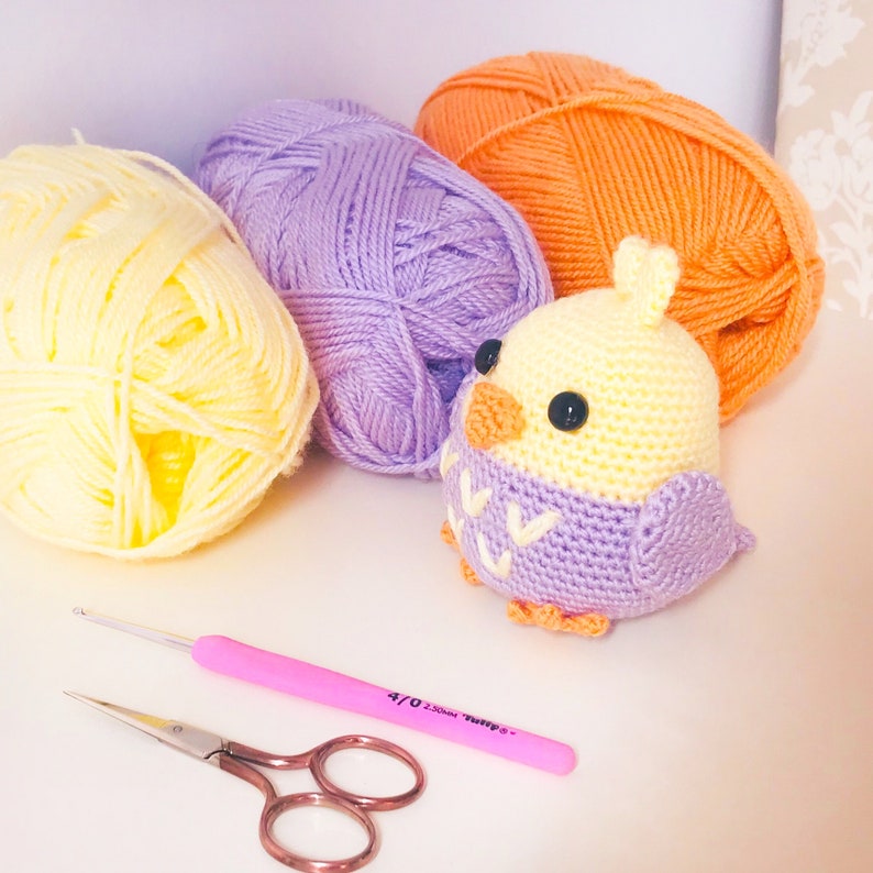 May include: A crocheted bird toy with yellow and purple yarn, along with three balls of yarn in yellow, purple, and orange. A pink crochet hook and a pair of scissors are also included.