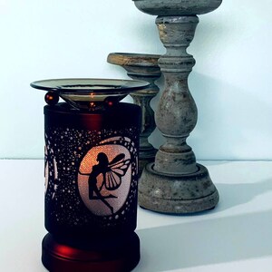 May include: A decorative, dark red electric oil warmer with a fairy silhouette design. The warmer has a glass dish and two red spheres. Two gray wooden candle holders are in the background.