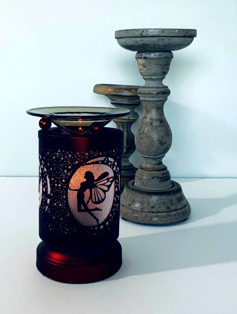 May include: A decorative, dark red electric oil warmer with a fairy silhouette design. The warmer has a glass dish and two red spheres. Two gray wooden candle holders are in the background.