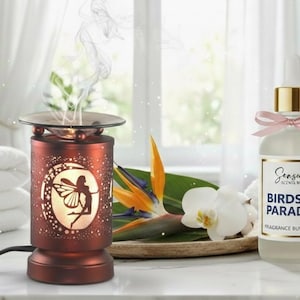 May include: A decorative aromatherapy oil diffuser with a fairy design, a bottle of fragrance burning oil, a lit candle, and white towels. The oil diffuser is red-brown and has a glass top. The oil bottle has a gold cap and a pink ribbon.