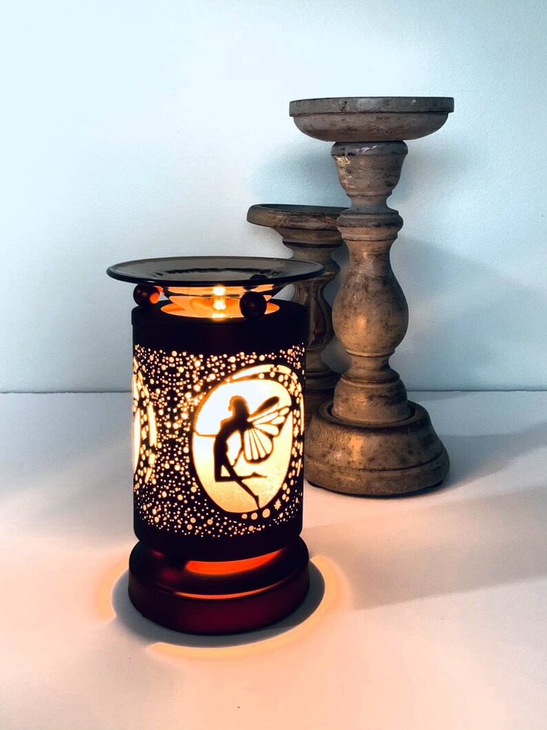 May include: A black electric wax warmer with a fairy silhouette design. The warmer has a round top and base, with a lit candle inside. Two wooden candle holders are in the background.