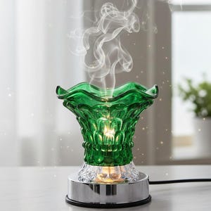 May include: A green glass essential oil diffuser with a decorative floral design. The diffuser has a chrome base and a clear, textured bottom. Wisps of white vapor rise from the top, suggesting the diffusion of essential oils. The diffuser is on a white surface.