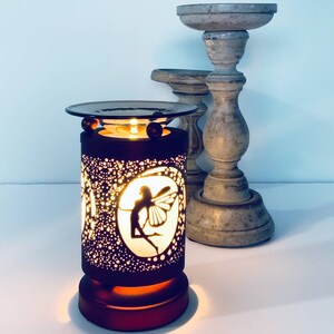 May include: A dark brown electric oil warmer with a fairy silhouette design. The warmer has a round top and base, with a cylindrical body perforated with small holes. Two wooden candle holders are in the background.