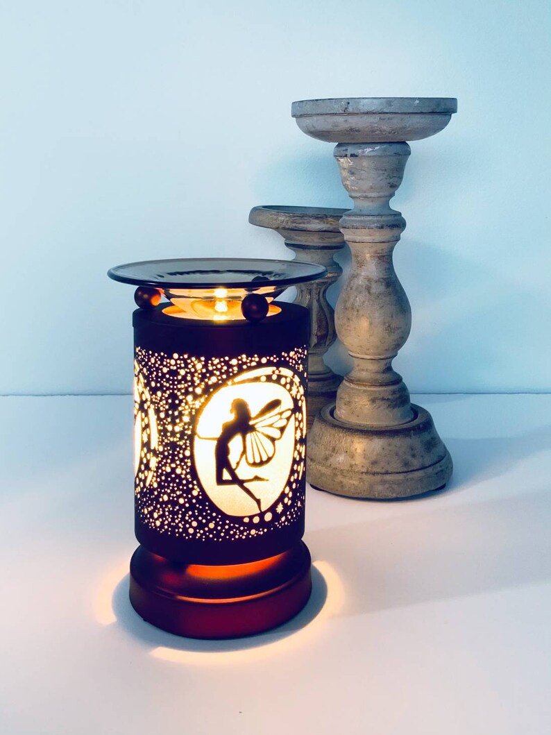 May include: A dark brown electric oil warmer with a fairy silhouette design. The warmer has a round top and base, with a cylindrical body perforated with small holes. Two wooden candle holders are in the background.