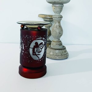May include: A red electric oil warmer with a fairy silhouette design. The warmer has a glass dish and two red decorative balls. Two decorative wooden candle holders are in the background.