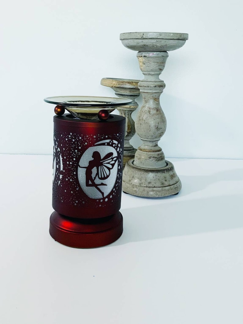 May include: A red electric oil warmer with a fairy silhouette design. The warmer has a glass dish and two red decorative balls. Two decorative wooden candle holders are in the background.