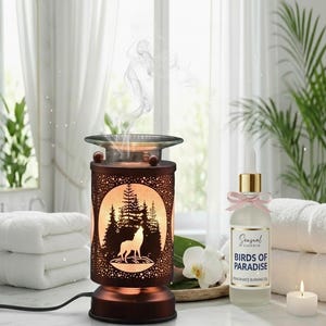 May include: A bronze-colored essential oil diffuser with a wolf and forest silhouette design. The diffuser is lit from within, and a bottle of "Birds of Paradise" fragrance oil sits nearby. A lit candle and towels complete the scene.
