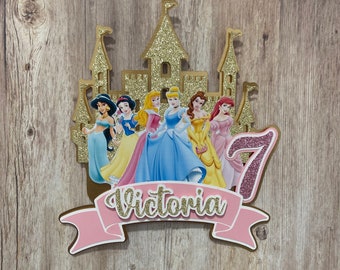 Pode incluir: Um topo de bolo de castelo rosa e dourado com sete princesas Disney, uma faixa rosa com o nome "Victoria" e o número "7" em glitter rosa.