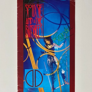May include: A promotional poster for Siouxsie and the Banshees, featuring a blue background with the band's name in red lettering. The image includes a band member and is signed. The poster has a red border.