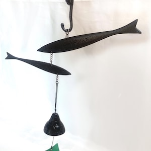 Può includere: Campanello eolico in metallo nero con due ornamenti a forma di pesce sospesi a un gancio decorativo. Una campana e un'etichetta con testo giapponese sono appese sotto. Il campanello eolico è tenuto in mano.