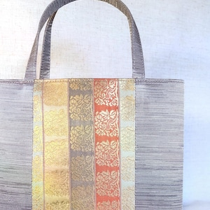 Könnte beinhalten: Eine rechteckige Tragetasche mit einem gestreiften grauen Körper und einem Mittelteil aus vertikalen Streifen in Gold, Grau und Koralle mit floralen Mustern. Die Tasche hat passende graue Griffe.