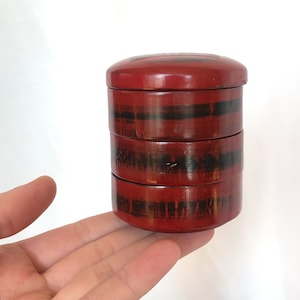 Puede incluir: Una caja bento de madera de color rojo y negro con tapa redonda. La caja está apilada en cuatro niveles y está sobre una mano.