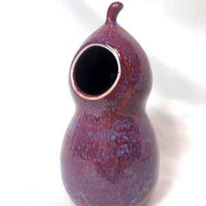 Puede incluir: Un pequeño jarrón de cerámica hecho a mano en un esmalte morado oscuro y azul. El jarrón tiene una forma redondeada única con una gran abertura en la parte superior.