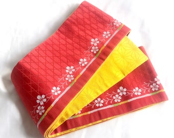 May include: A red and yellow fabric sash with a white floral pattern. The sash is folded in thirds and has a diamond pattern on the red fabric.