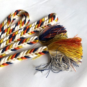 Può includere: Un cordoncino intrecciato con nappe di colore blu navy, bianco, arancio e giallo. Il cordoncino è realizzato in un materiale spesso e tessuto e ha una nappa a ciascuna estremità.