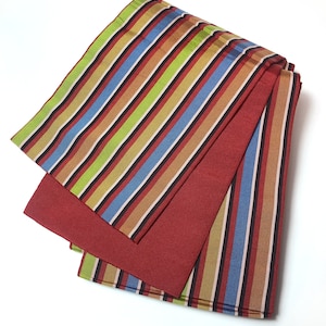 Puede incluir: Una tela de rayas rojas y multicolores con una tela roja debajo. La tela está doblada y tiene un patrón de rayas con rayas rojas, azules, verdes, amarillas y marrones.