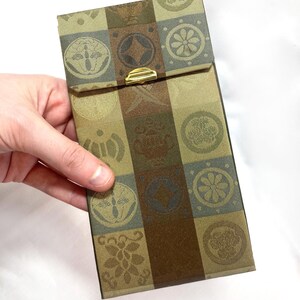 Pode incluir: Estojo retangular com um padrão geométrico repetido em tons de verde, castanho e dourado. O estojo tem um fecho dourado. O design inclui motivos florais e circulares. O estojo é segurado por uma mão.