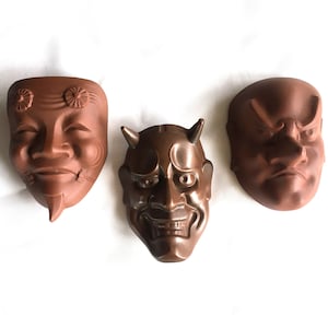 Peut inclure: Trois masques Noh japonais traditionnels en terre cuite, avec des expressions différentes. Les masques sont de couleur brune et présentent des détails complexes. Le masque de gauche a une expression souriante, le masque du milieu a une expression féroce et le masque de droite a une expression renfrognée.