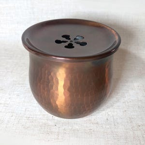 May include: A copper-colored, hammered metal container with a lid. The lid has a flower-shaped ventilation pattern. The container has a rounded shape and a textured surface, reflecting light. The overall design suggests a decorative or functional item.