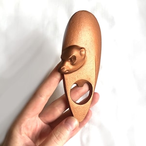 May include: A copper-colored bottle opener shaped like a meerkat. The meerkat's head is facing to the left, and the bottle opener has a cut-out for opening bottles.