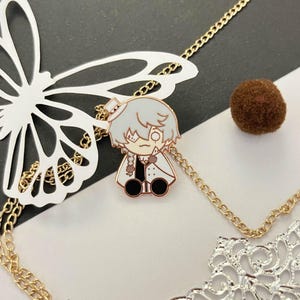 Peut inclure: Un accessoire décoratif en forme de papillon avec une chaîne dorée, une petite épingle de personnage de dessin animé et un coin en filigrane argenté. Un petit pompon marron est également présent. Le fond est blanc et noir.