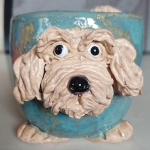 May include: A ceramic mug shaped like a dog with a light blue glaze. The dog has a light brown face with a black nose and white eyes.