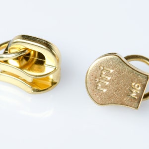 Puede incluir: Dos tiradores de cremallera dorados. Uno está en posición cerrada, mostrando los dientes entrelazados. El otro tirador está etiquetado como "riri" y "M6". Ambos tiradores tienen un anillo para la fijación.