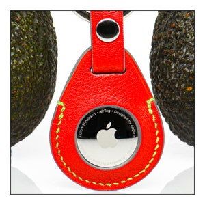 May include: Red leather AirTag holder with a green stitched border. The back of the holder features a silver Apple logo and the text "Ultra Wideband - AirTag - Designed by Apple in California - Assembled in China".