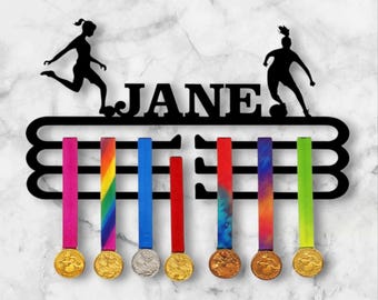 May include: Black metal medal holder with the name "JANE" and soccer player silhouettes. The holder displays medals with colourful ribbons. The ribbons are pink, rainbow, blue, red, tie-dye, and green. The medals are gold and silver.