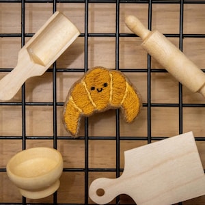 Könnte beinhalten: Eine Sammlung von Holzbackwerkzeugen und ein lächelndes, gesticktes Croissant. Die Werkzeuge umfassen eine Schaufel, eine Teigrolle, eine Schüssel und ein Schneidebrett. Die Artikel sind auf einem schwarzen Gitter vor einem hellbraunen Hintergrund angeordnet, was ein Bäckereithema hervorruft.