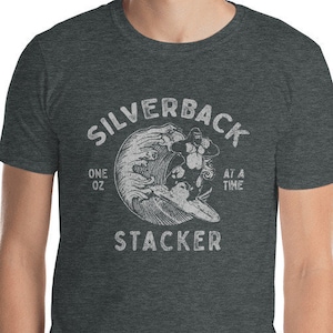 Peut inclure: Un t-shirt gris foncé chiné avec un motif blanc d'un gorille sur une vague. Le motif comprend le texte "Silverback Stacker" et "One Oz At A Time".