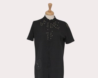 May include: A black jumpsuit with short sleeves and a zip closure. The jumpsuit has a small pocket on the left leg and gold-coloured accents on the neckline and shoulders.