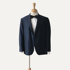 May include: A dark blue suit jacket with a black satin lapel and a white shirt with a black bow tie. The jacket is on a wooden mannequin.