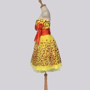 May include: A yellow tulle dress with a red satin bow and gold and orange sequins. The dress is on a wooden mannequin.